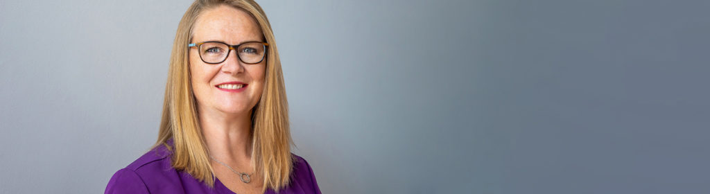 blonde lady wearing purple blouse and spectacles smiling at camera